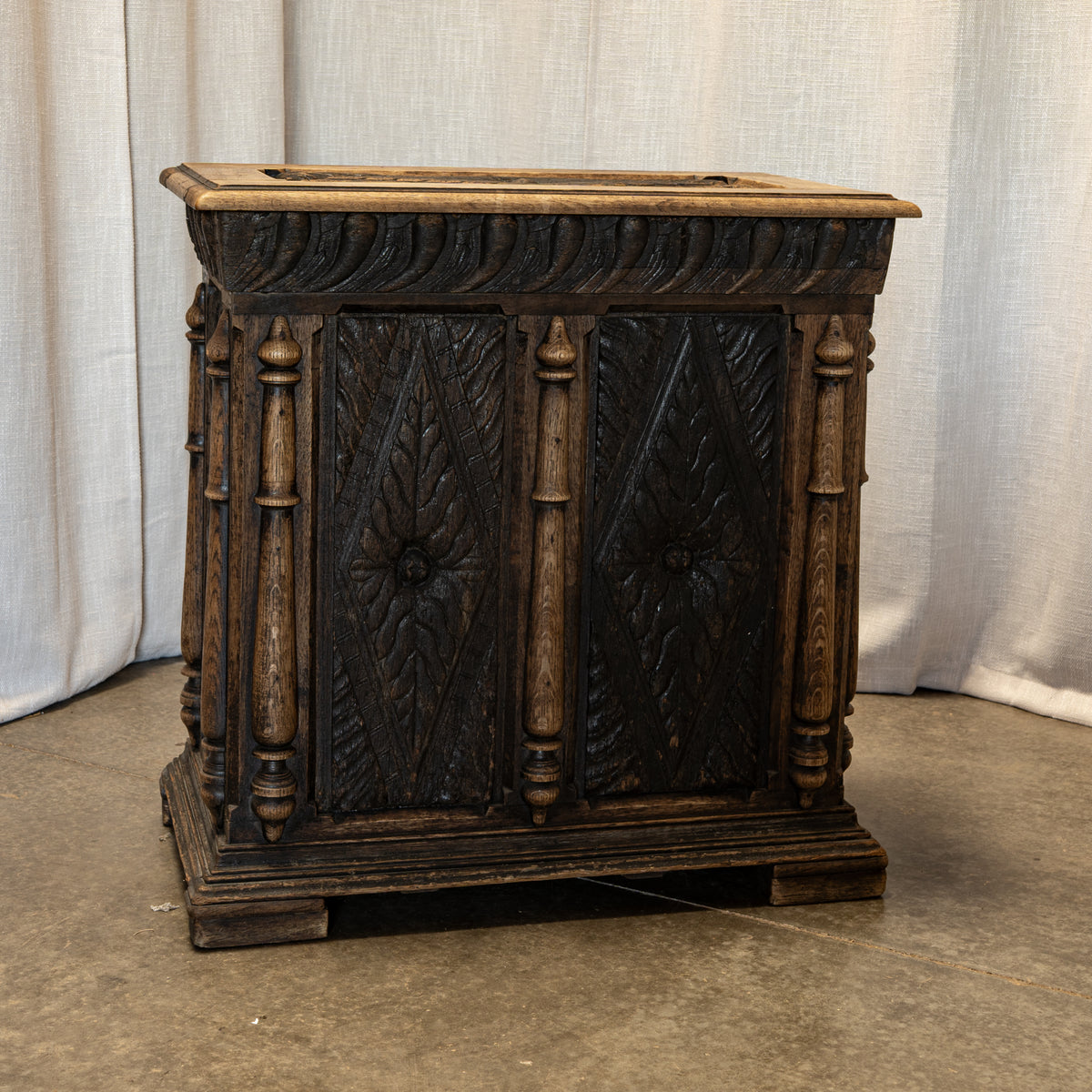 An ornate dark carved wooden chest or cabinet with decorative panel doors and turned side posts, photographed indoors against a light fabric backdrop and concrete floor.