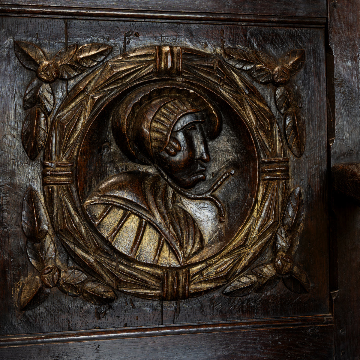 Close-up of an ornate carved wooden panel featuring a circular portrait bust in relief, surrounded by a laurel-like wreath and leaf motifs, in dark brown wood with warm golden highlights.