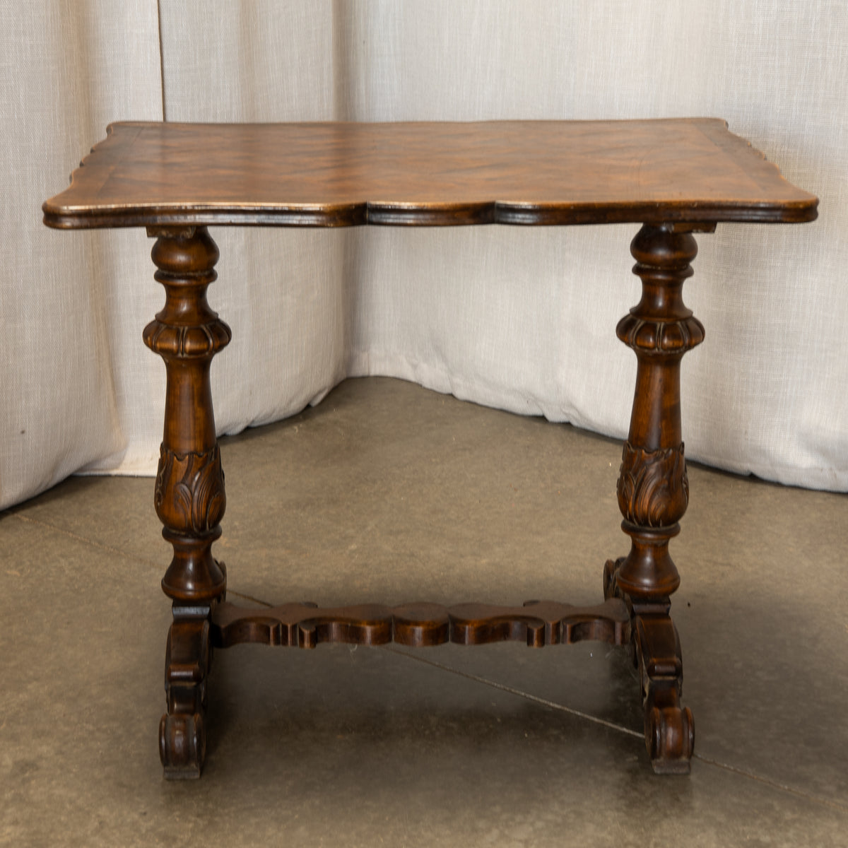 An antique-style wooden end table with a scalloped top, turned carved legs, and a stretcher base, shown against a neutral draped fabric backdrop.