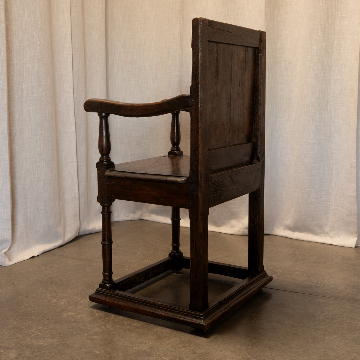 A dark brown wooden armchair with a high paneled backrest, turned legs, and a square footrest, photographed against a draped off-white fabric backdrop.