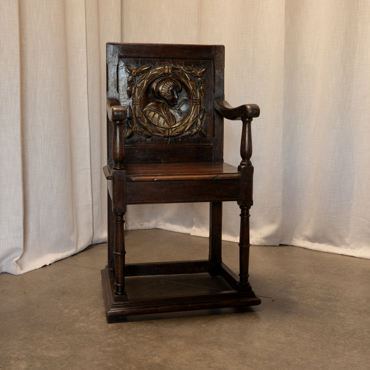 A dark brown carved wooden armchair with a square backrest featuring an ornate relief carving, set against a neutral beige fabric backdrop.
