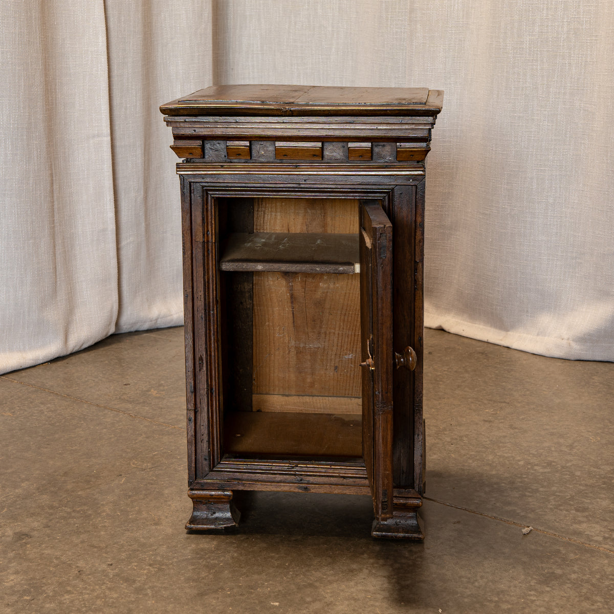 A small antique dark wood cabinet with one door open, showing an interior shelf, set against a neutral draped fabric backdrop.