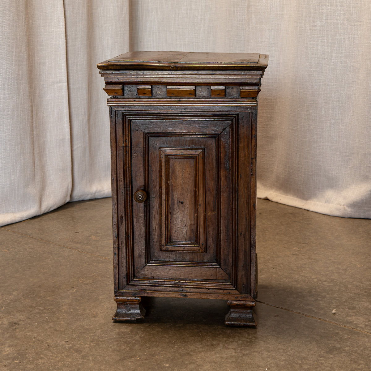 A dark brown wooden nightstand with a single paneled door, decorative carved top, and turned feet, set against a neutral draped fabric backdrop.