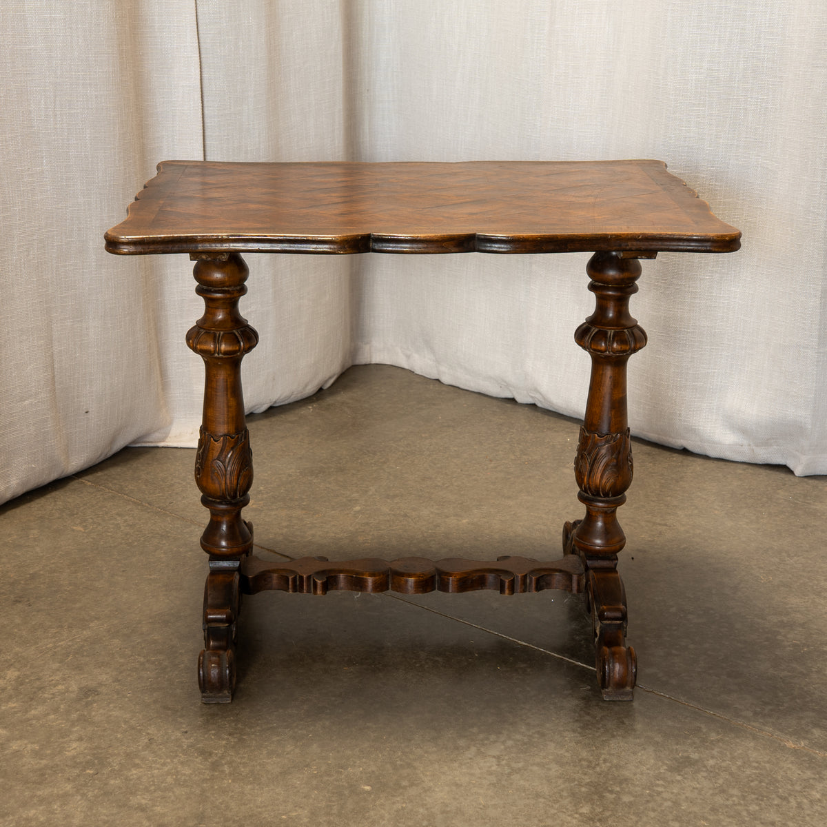 An antique dark brown wooden table with carved turned legs and a scalloped apron stretcher, set against a neutral draped fabric backdrop.
