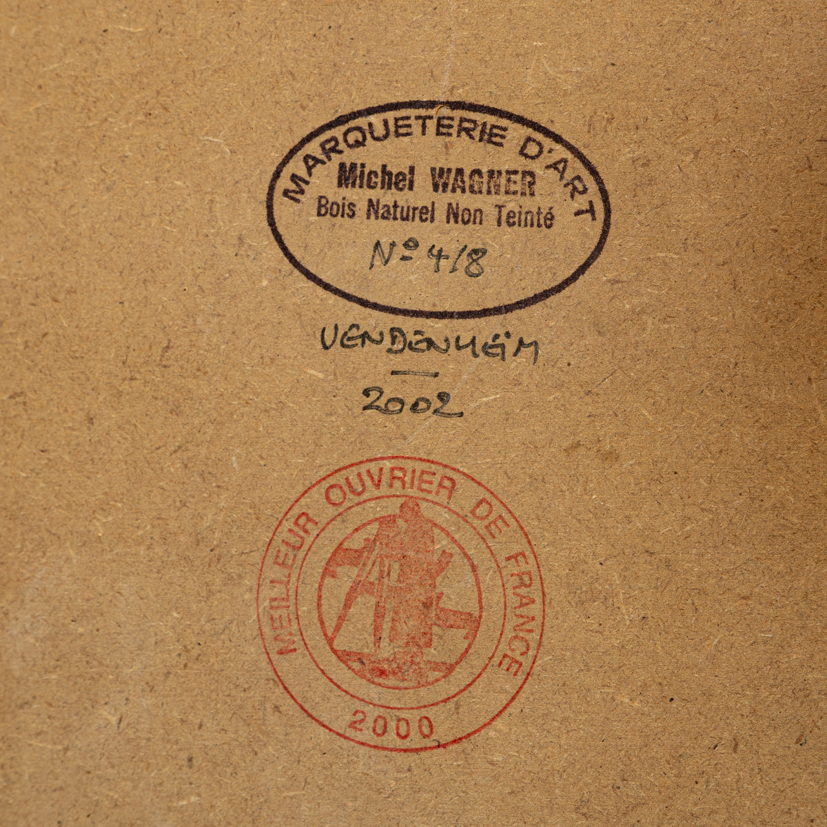 A close-up of a textured brown paper surface with two stamped marks: a black oval stamp reading 'MARQUETERIE D'ART Michel WAGNER Bois Naturel Non Teinté N°4/8 Venduém 2002' and a red circular stamp below it.
