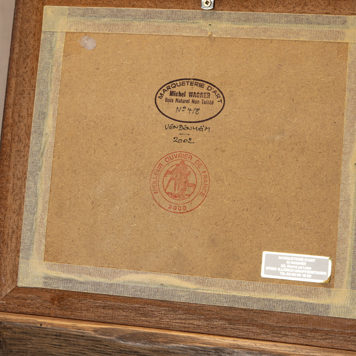 The back of a framed artwork or print, showing a brown cardboard backing with a wooden frame, tape border, a small product label, and two stamps including one circular black stamp reading Michel Braquet and one red circular stamp.