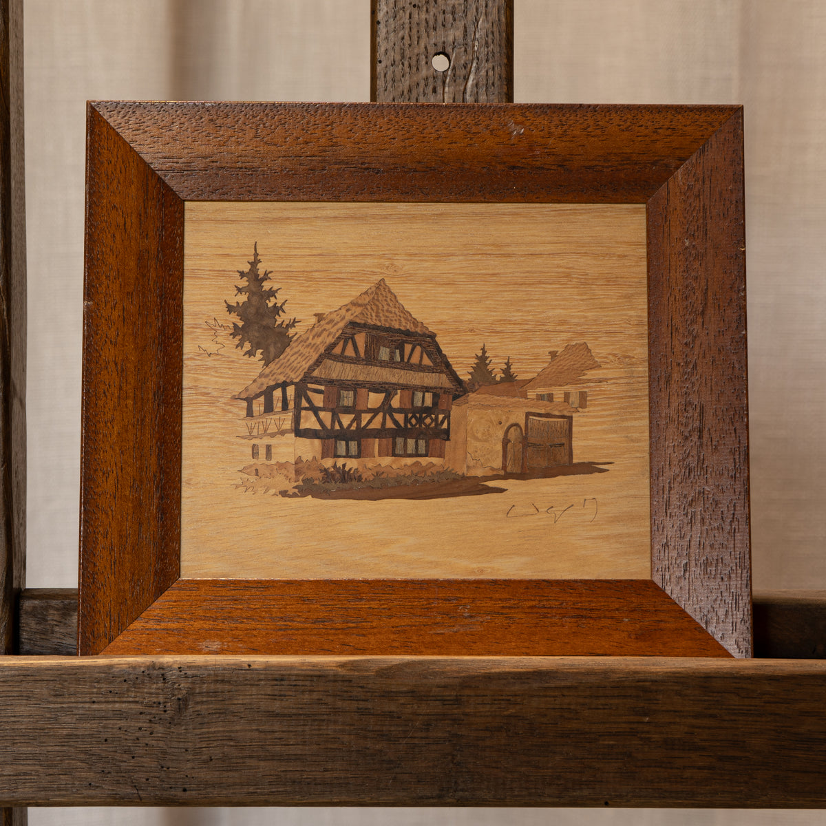 A square wooden framed artwork featuring an engraved or burned rustic half-timber house scene with trees and a small cart, displayed on a wooden stand against a light neutral background.