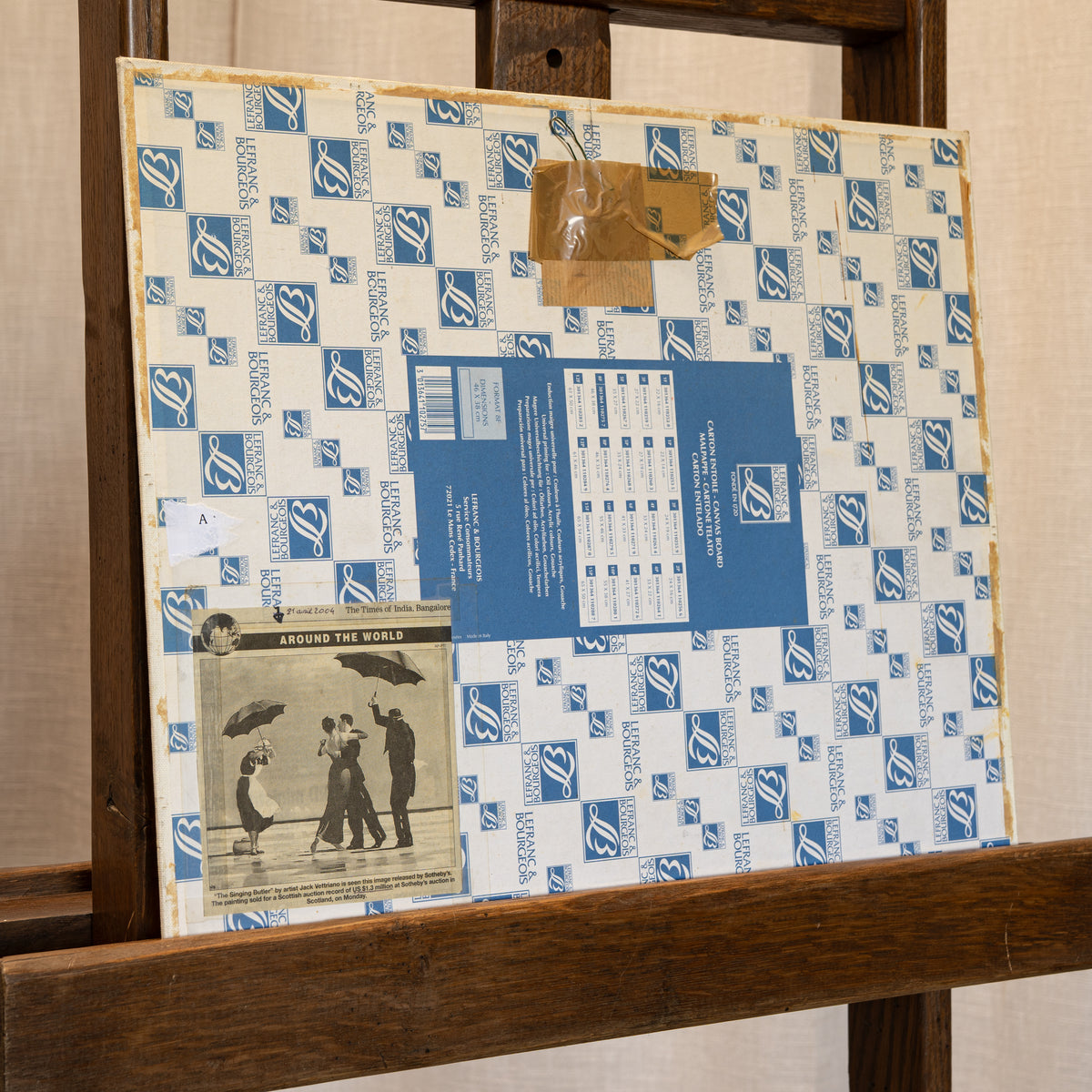 A square music record sleeve or album cover wrapped in protective blue-and-white branded paper, photographed resting on a wooden display stand against a beige wall.