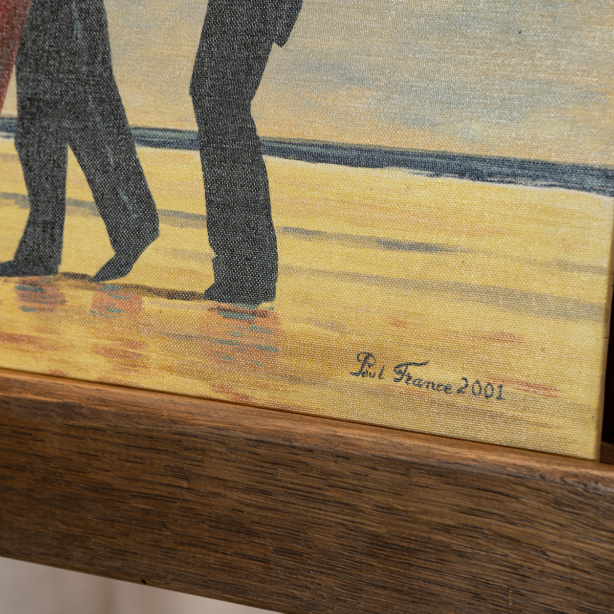 Close-up of a textured oil painting showing the lower legs of two people walking on a sandy beach, with a dark wooden easel visible in the foreground and the signature 'Eli France 2001' on the canvas.