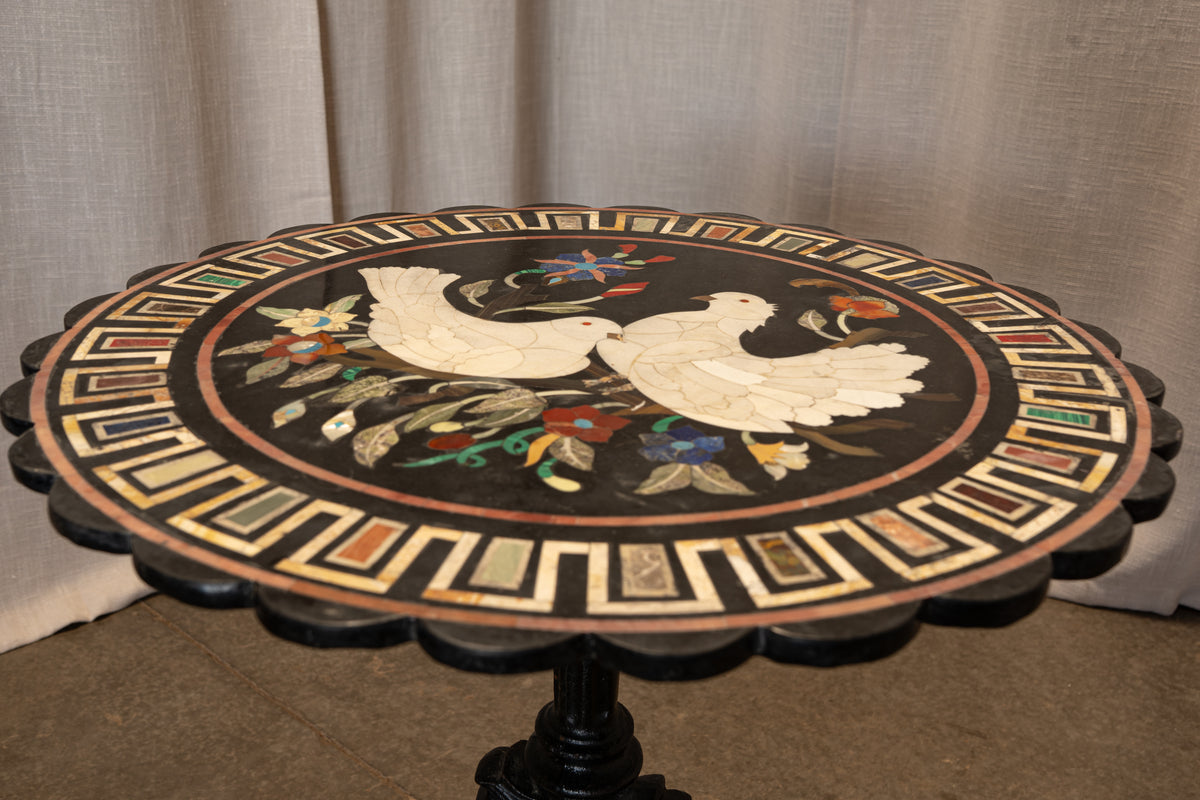 A round pedestal table with a scalloped black edge and an intricate inlay design. The top features a black center with two white doves, colorful floral motifs, and a decorative border of geometric inlays, set against a neutral beige curtain background.
