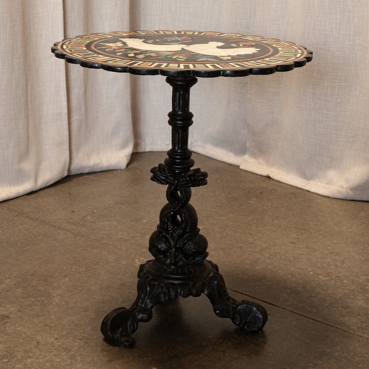 An ornate black cast iron pedestal table with a scalloped mosaic top featuring a white dove and gold decorative border, set against draped beige fabric and a concrete floor.