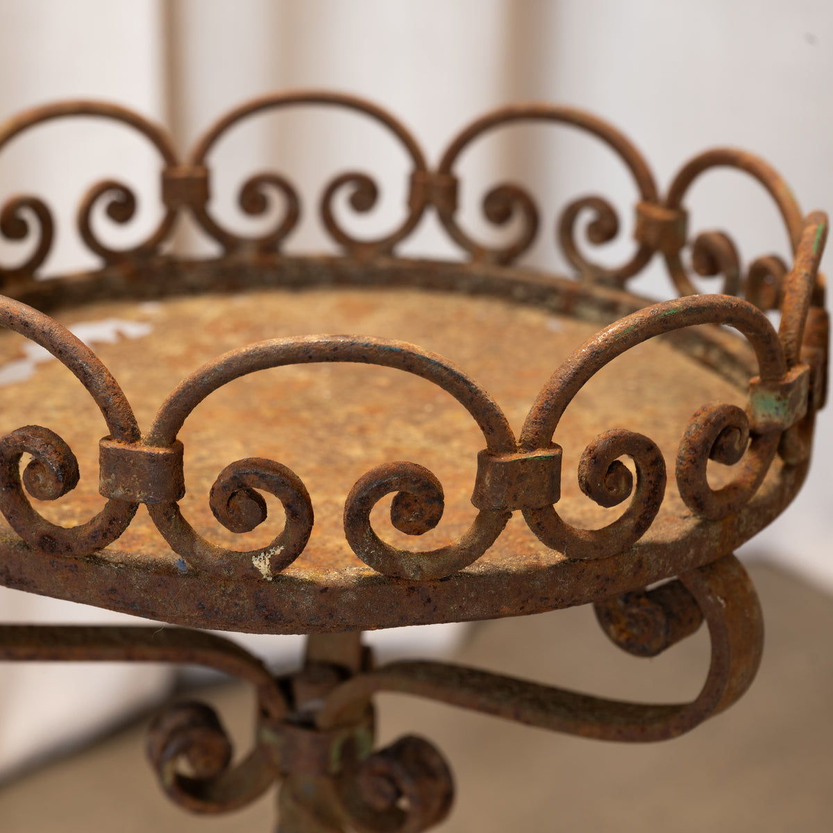 Close-up of a rustic wrought iron stand with a rusted brown finish and decorative scrollwork, showing the circular tray top and part of the pedestal base against a soft neutral background.