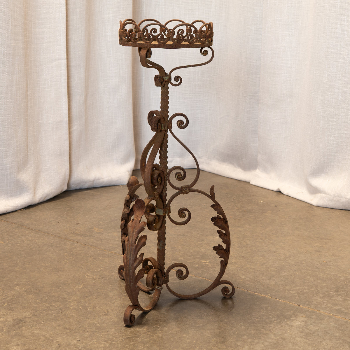 An ornate wrought iron candle holder stand with a crown-shaped top, scrolled details, and leaf-like flourishes, photographed against draped white fabric and a concrete floor.
