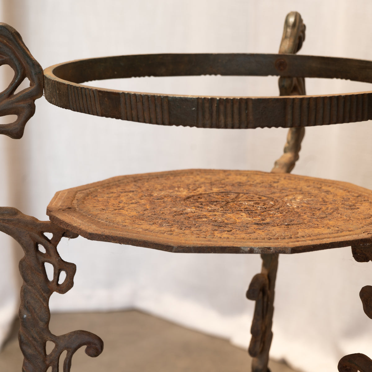An antique cast iron two-tier serving stand with ornate scrollwork legs and a round top tier, shown against a neutral draped backdrop.
