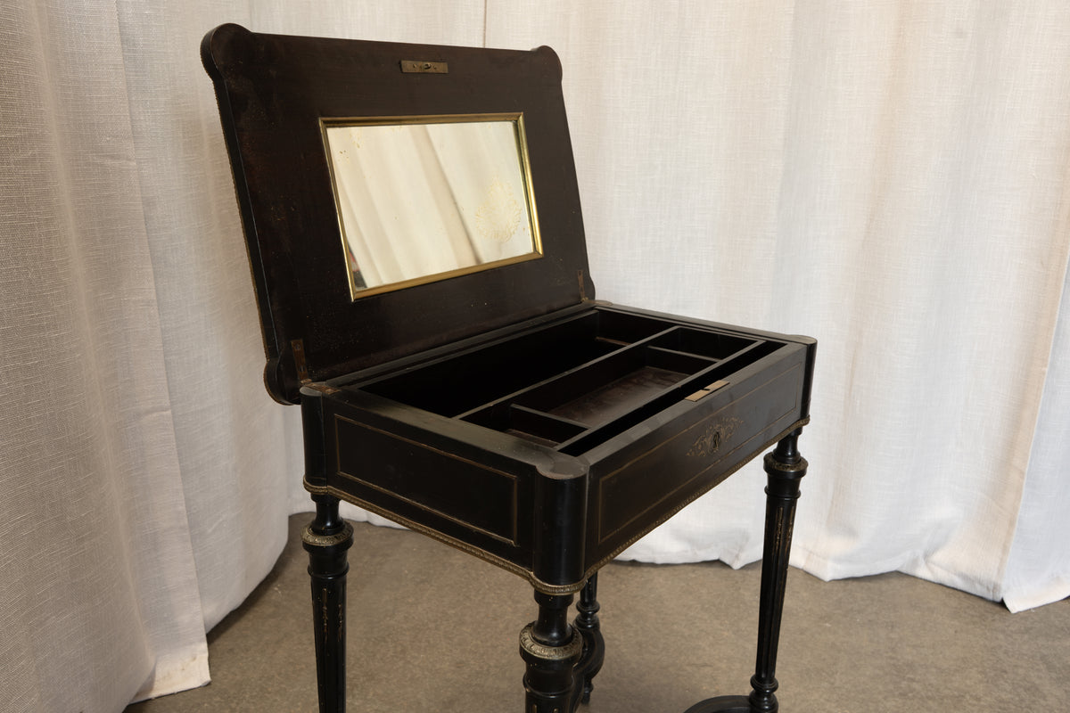 An antique black lacquered vanity desk with an open lid showing a mirror and interior compartments, set against a white draped fabric backdrop.