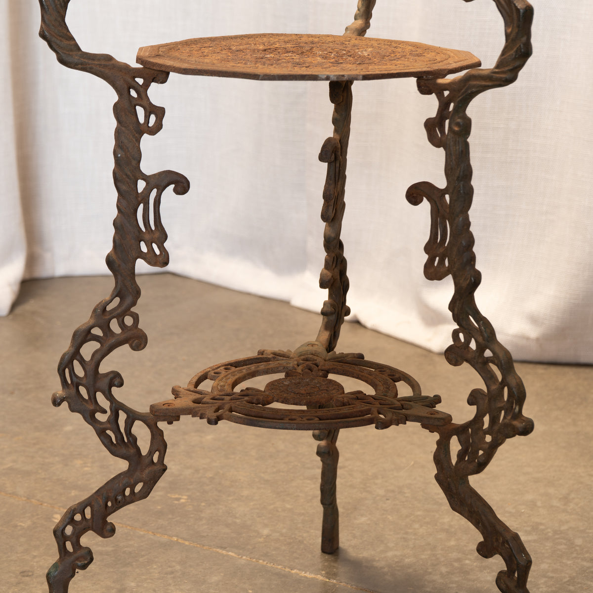 An ornate cast iron plant stand with a rusted brown finish, featuring a top tiered platform, a middle shelf, and a decorative backrest, photographed against a draped white fabric backdrop.