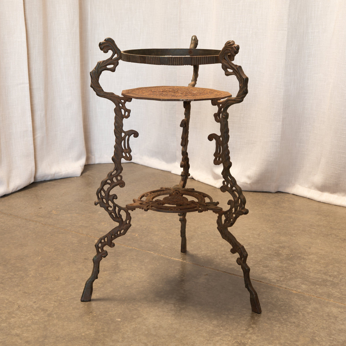 An ornate vintage cast iron stand with a rustic dark brown finish and a light brown woven rattan top and middle shelf, set against a draped white curtain backdrop.