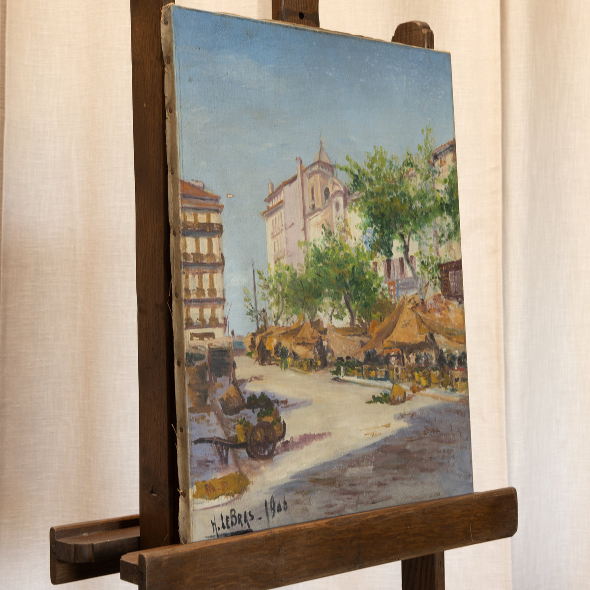 An oil painting of a street scene with buildings, trees, and market stalls, displayed on a wooden easel against a beige curtain background.