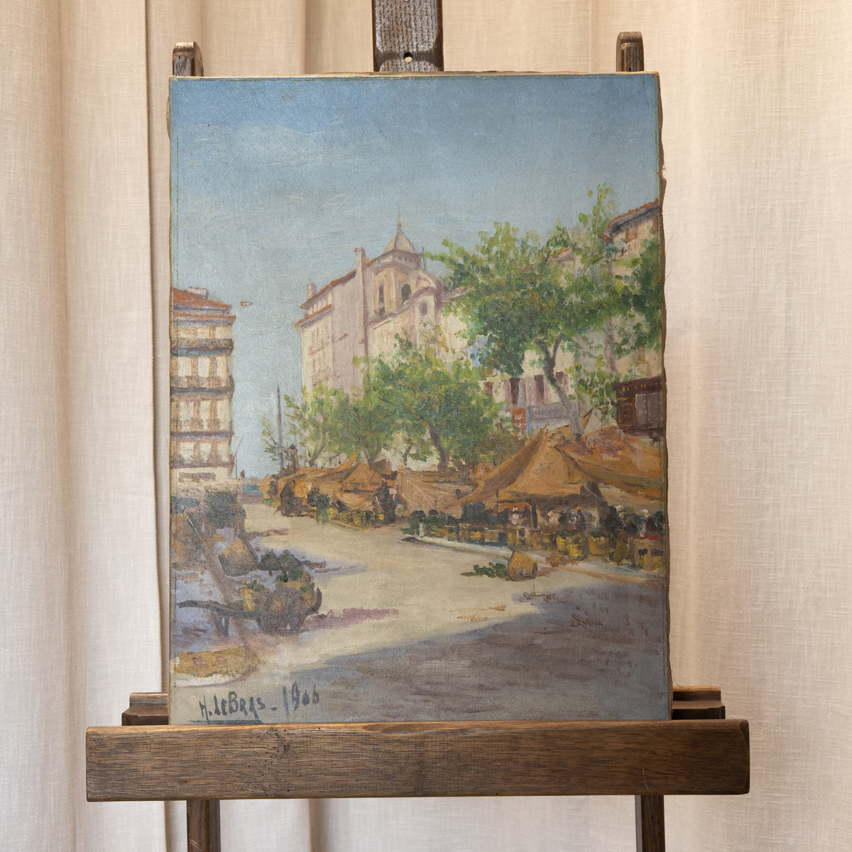 An oil painting of a European street scene with cafes, trees, and buildings, displayed on a wooden easel against a beige curtain background.