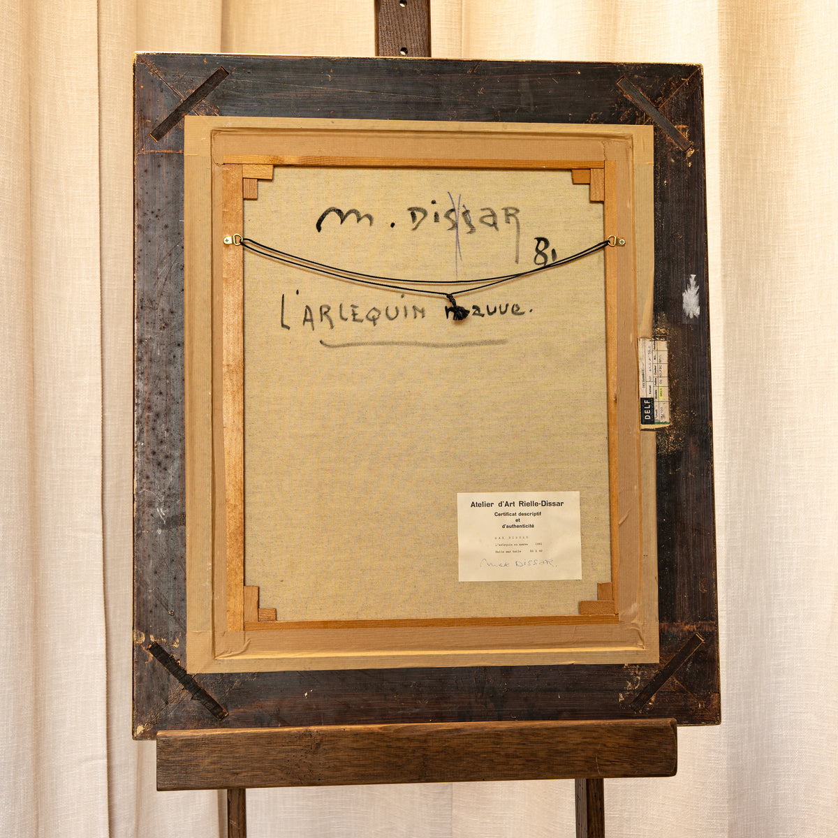 The back of a framed artwork on an easel, showing a beige backing board with handwritten text, a metal wire support, and a small paper label, set against a cream fabric backdrop.