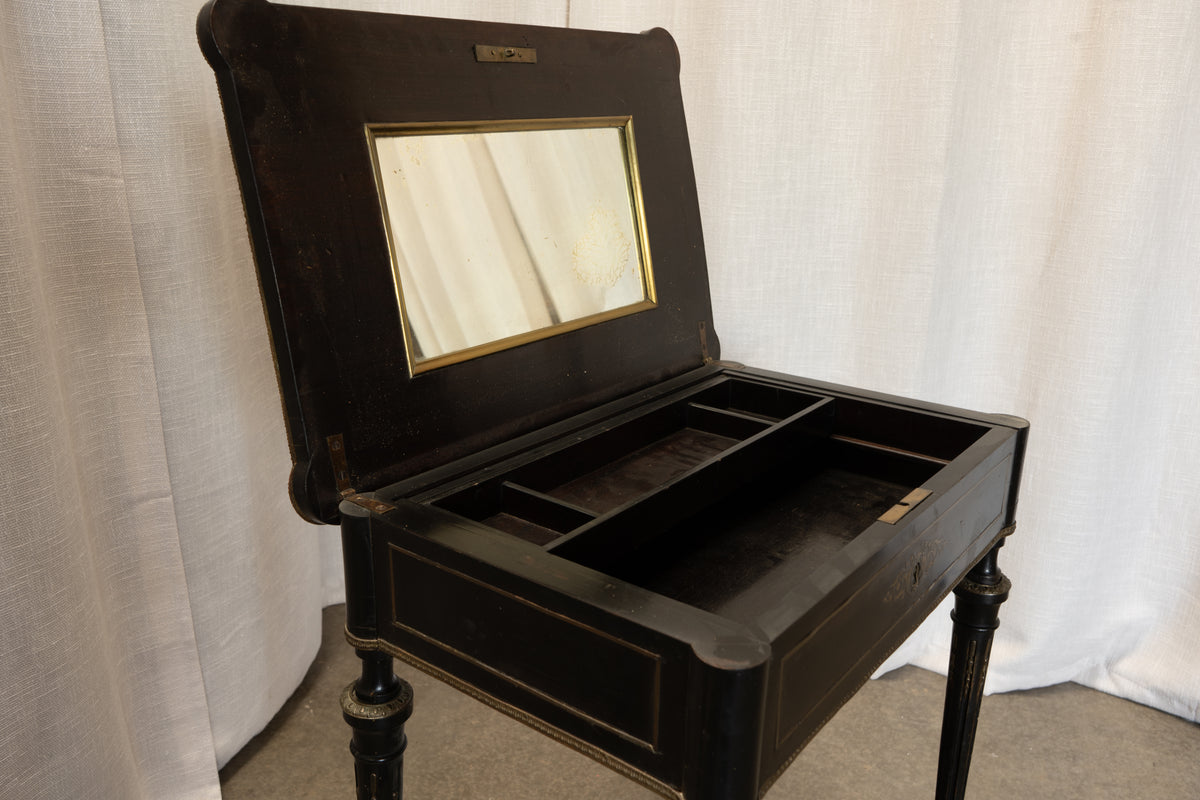 An antique black wooden dressing table with an open hinged lid containing a mirror and interior compartments, set against a white curtain backdrop.