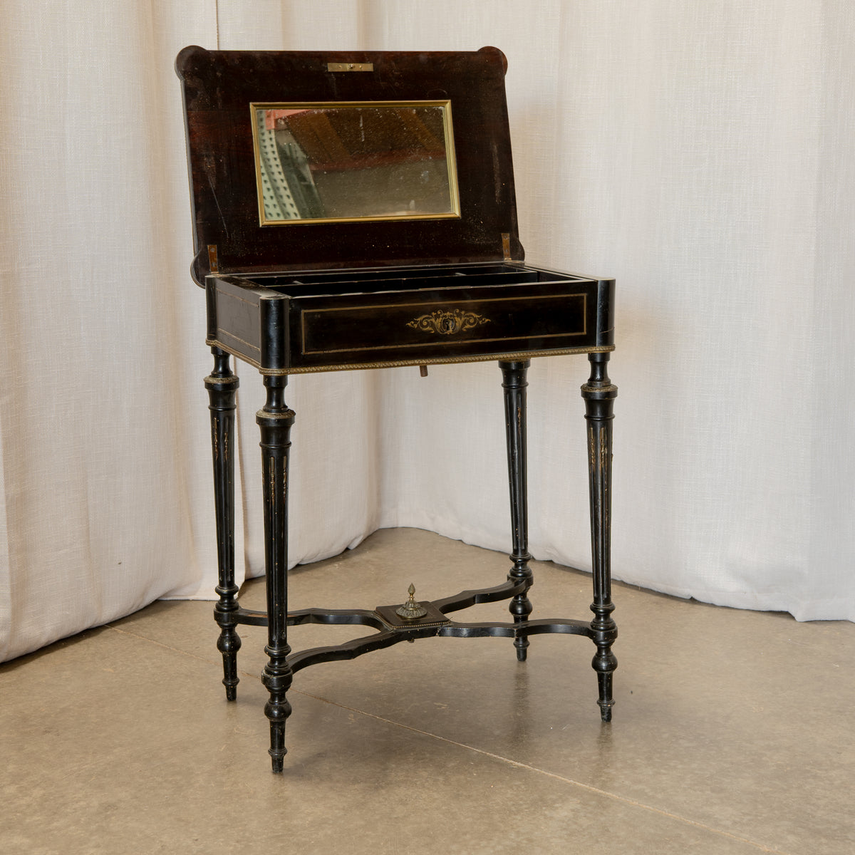 An antique black lacquered writing desk with a hinged wooden back panel, an inset mirror, and a single drawer with gold-tone hardware, set against a draped white fabric backdrop.