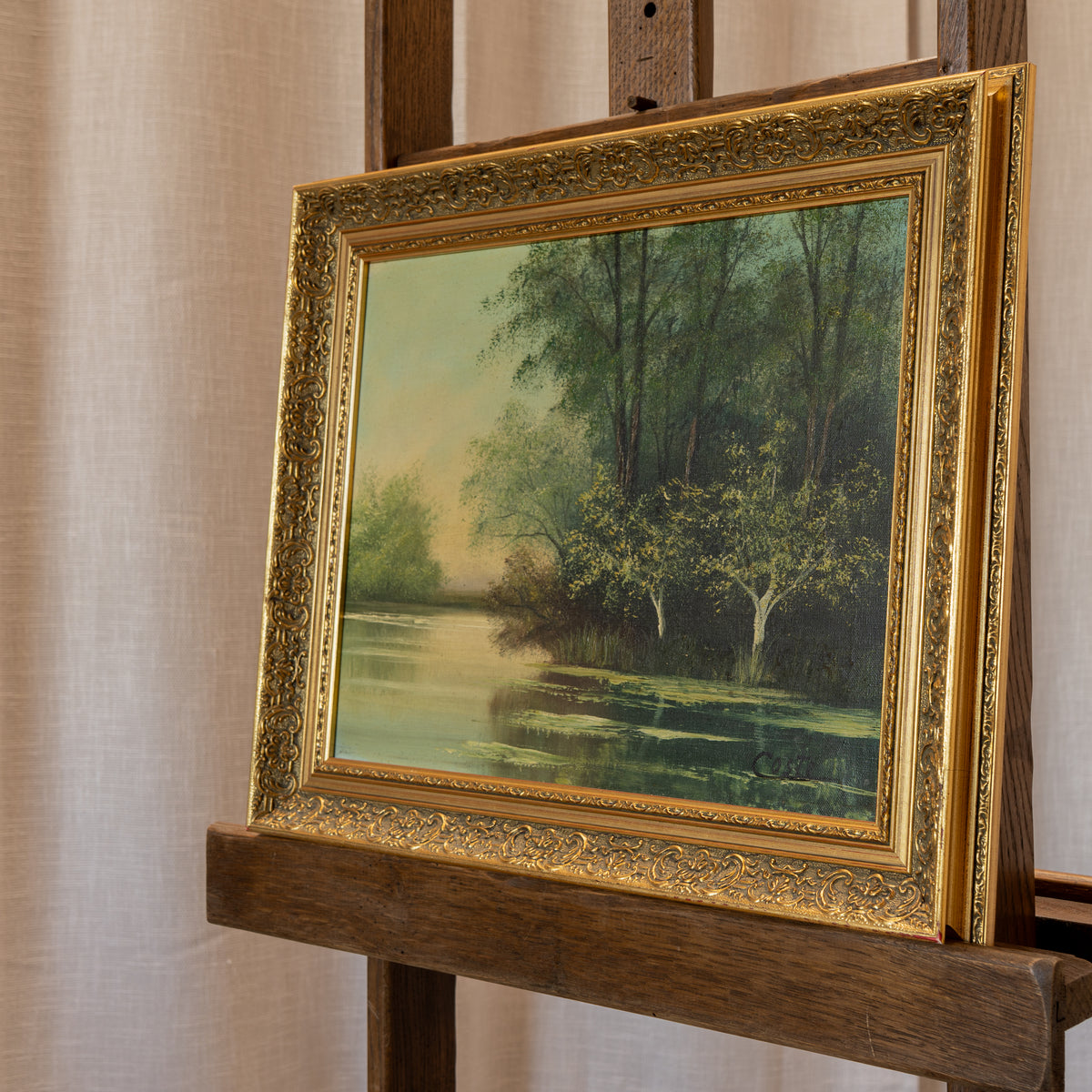 A framed landscape oil painting of a tree-lined river, featuring a detailed ornate gold frame, displayed on a wooden easel against a neutral beige curtain background.