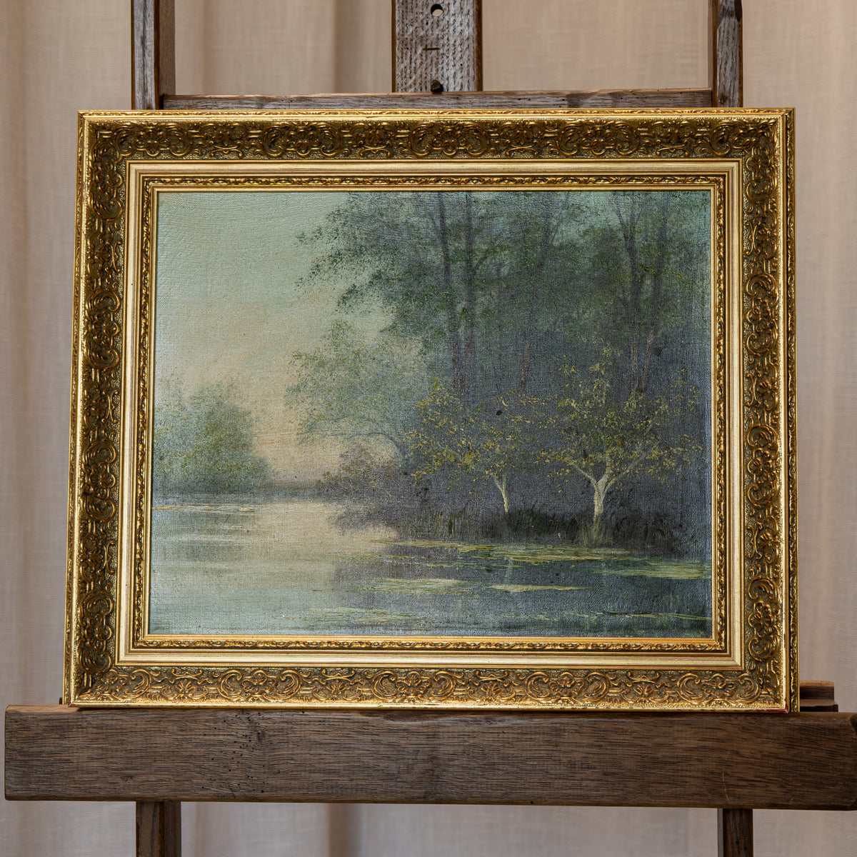 A framed landscape painting of a tree-lined pond, displayed on a wooden easel. The artwork features a soft, impressionistic scene with green and blue tones, enclosed in an ornate gold frame.