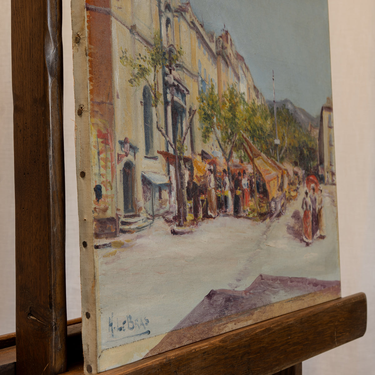 An oil painting on canvas depicting a European street scene with market stalls, figures, and buildings, mounted on a wooden easel.