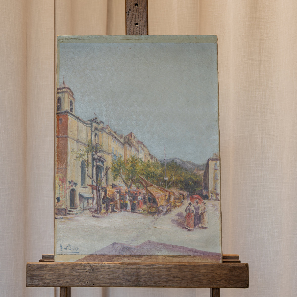 An oil painting of a European street scene with buildings, market stalls, and pedestrians, displayed on a wooden easel against a beige curtain background.