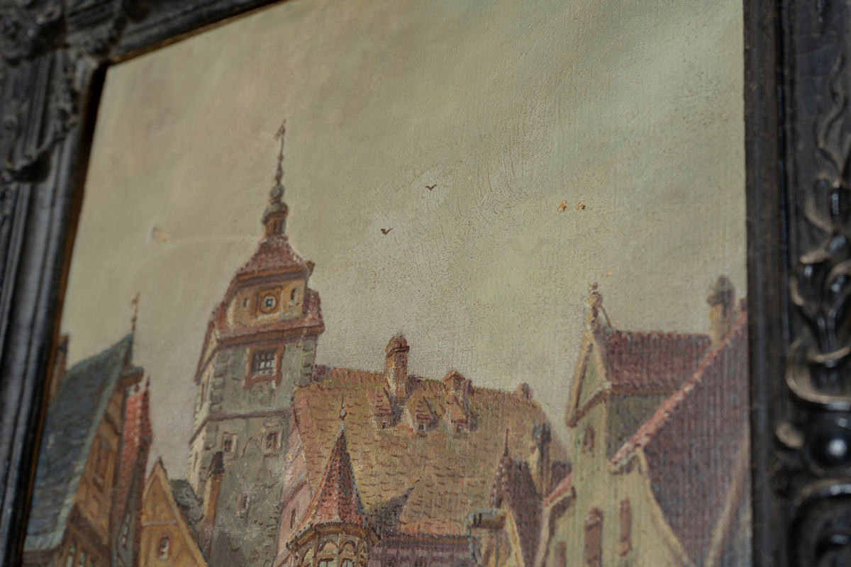 A close-up of a framed painting depicting a European town with a prominent clock tower, warm brown rooftops, and a pale sky. The ornate dark frame is visible on the left and right edges.