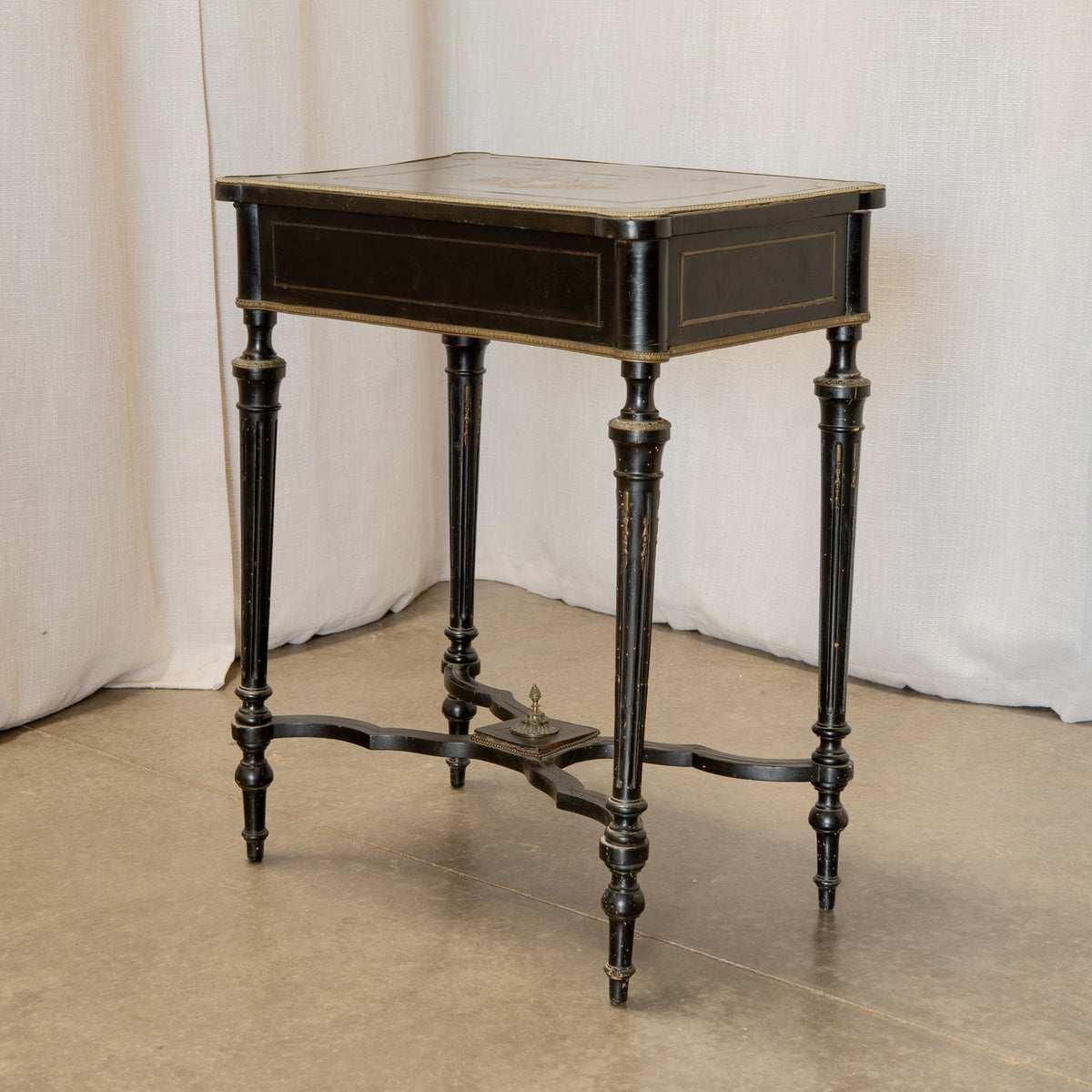 A vintage black wooden writing desk or side table with turned legs, a brass foot pedal, and gold-toned trim, set against a draped white fabric backdrop.