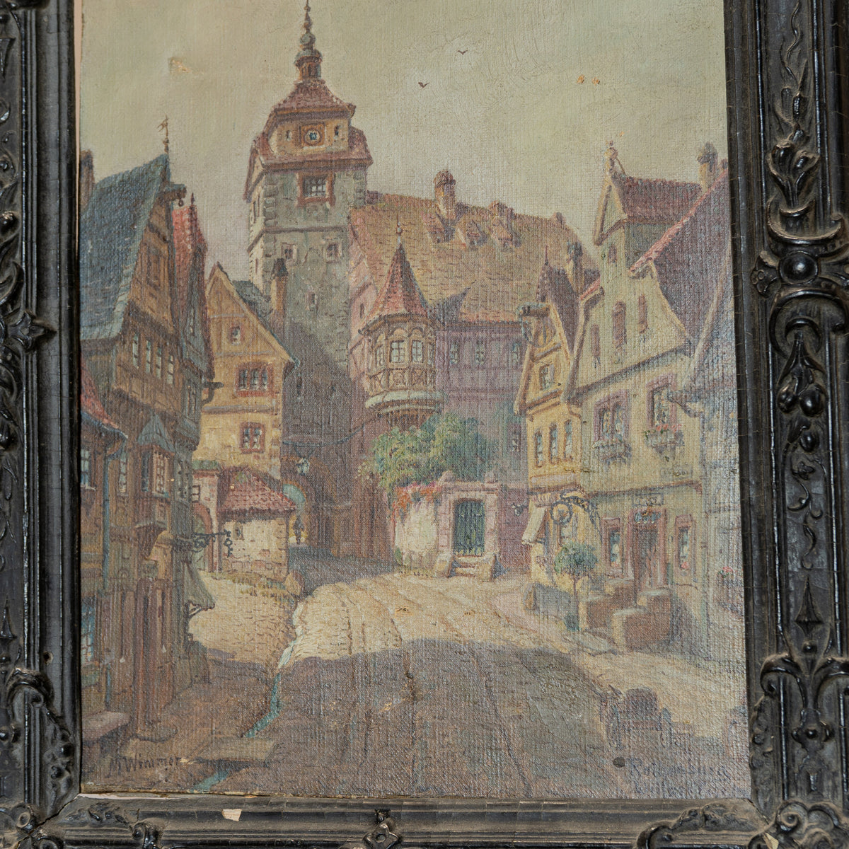 An antique oil painting of a European town street with half-timbered houses and a tower, housed in an ornate dark carved wooden frame.
