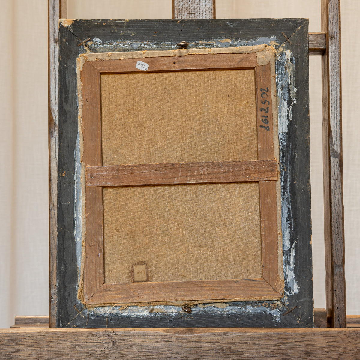 The back of a vintage wooden framed artwork, showing the canvas backing, distressed dark gray paint, and wooden frame, displayed on a wooden stand against a neutral cream curtain background.