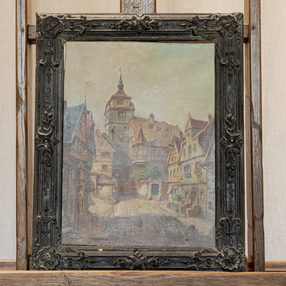 A framed painting of a European town street scene with historic buildings and a tower, displayed on a rustic wooden easel against a cream curtain backdrop.