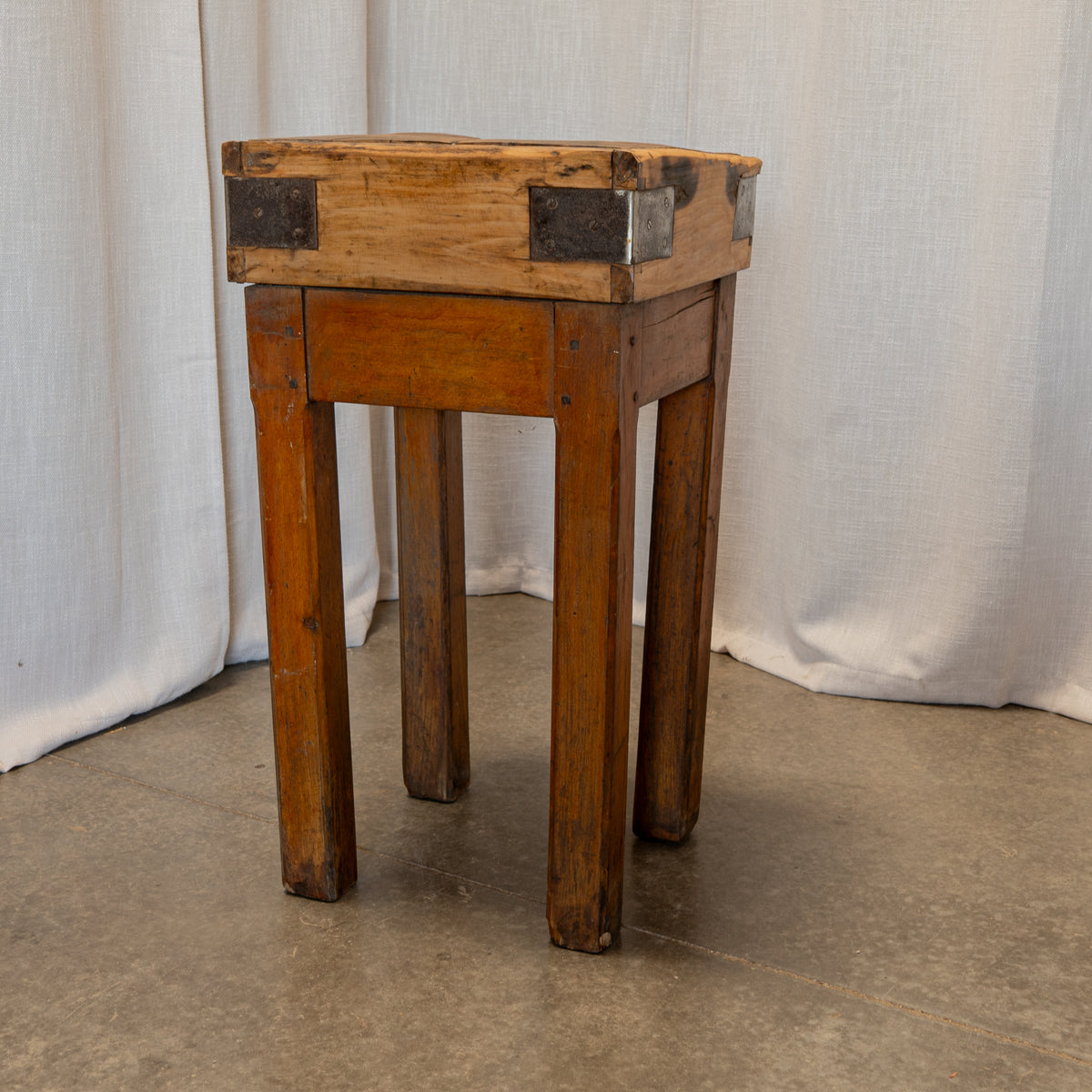A rustic wooden stool with a square top, single drawer, and four legs, featuring dark metal corner brackets, set against a draped white fabric backdrop.