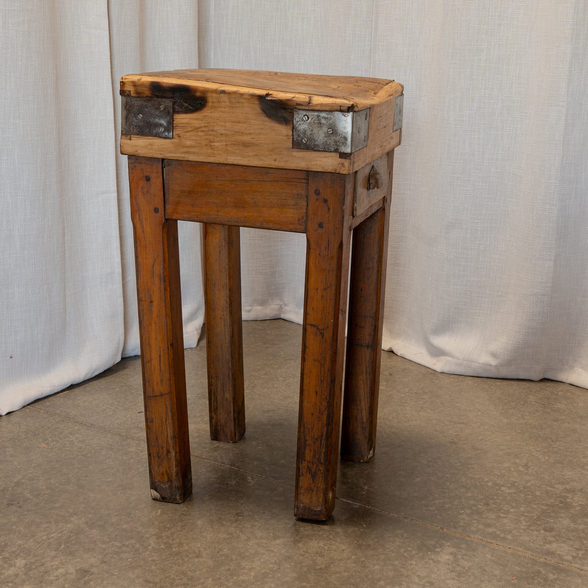A rustic wooden stool with a thick top, four legs, and dark metal corner brackets, set against a draped white fabric backdrop.