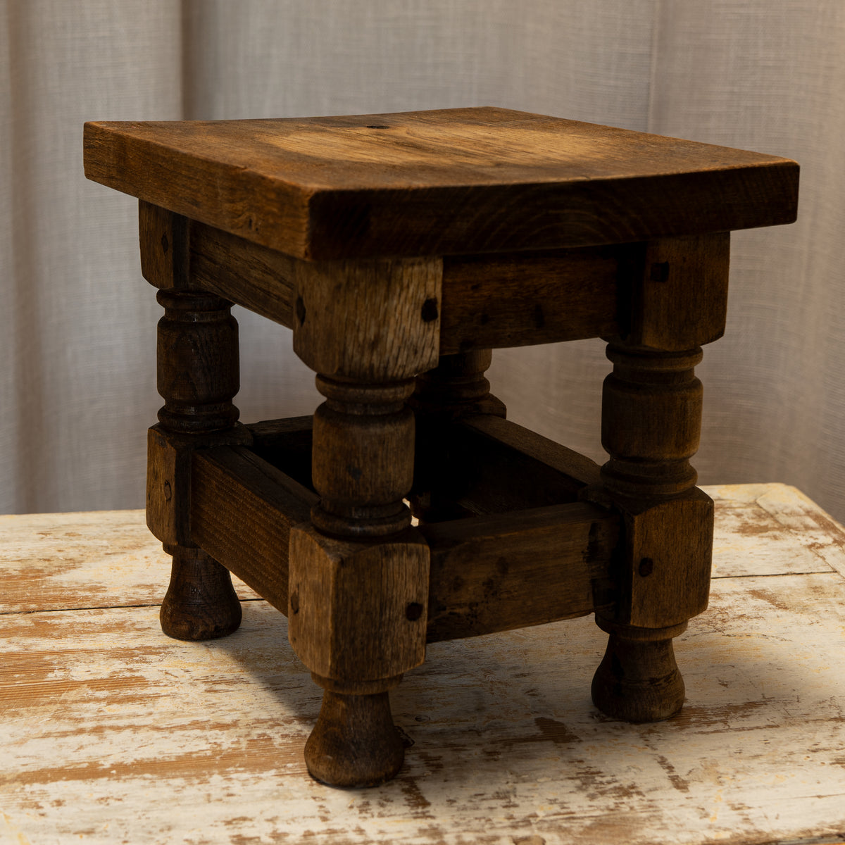A rustic dark brown wooden stool with turned legs and a square seat, shown on a distressed white wooden surface against a neutral fabric backdrop.