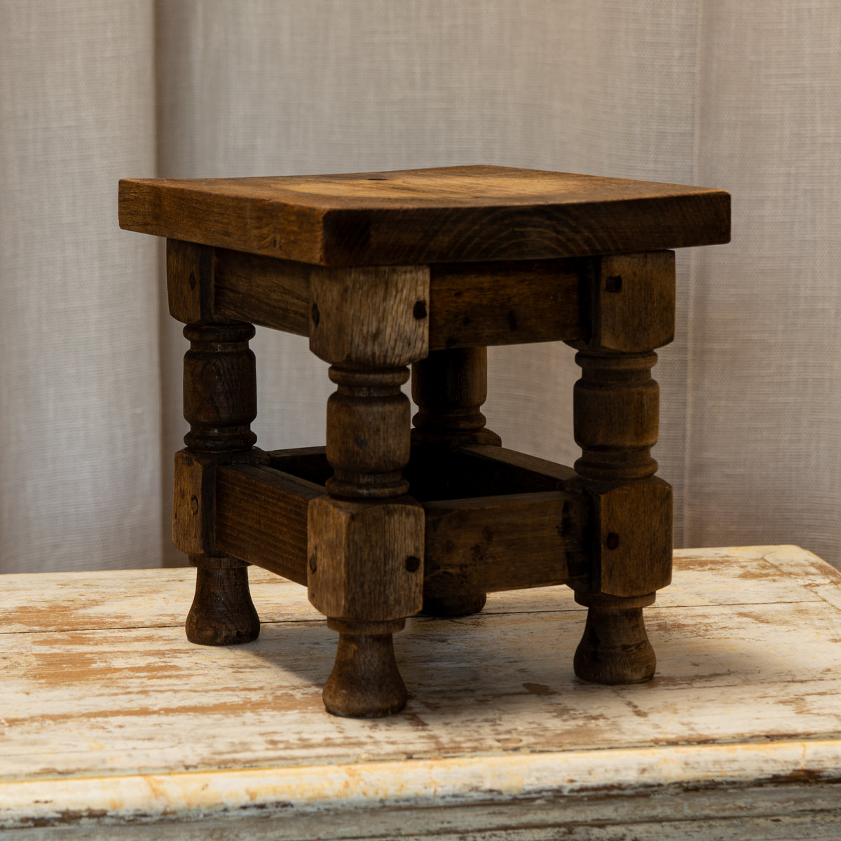 A rustic dark brown wooden stool with turned legs and a square footrest, set on a distressed white wooden surface against a neutral beige fabric backdrop.