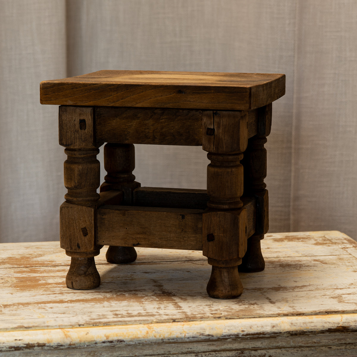 A small rustic wooden stool with turned legs and a thick square seat, shown on a distressed white wooden surface against a neutral beige fabric backdrop.