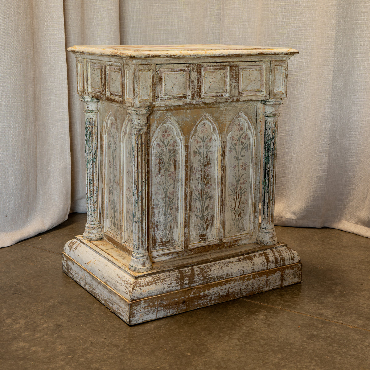 A distressed, vintage-style wooden altar or lectern with Gothic arched panel details and a weathered white and beige finish, set against a neutral draped fabric backdrop.