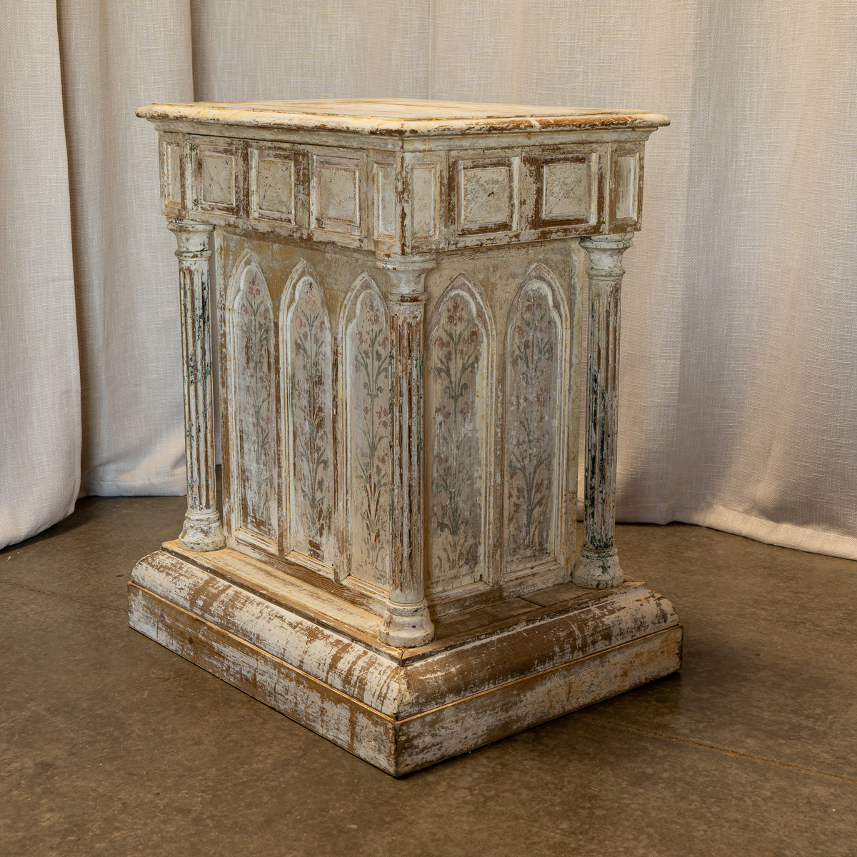 A distressed Gothic-style stone pedestal or lectern with arched panel details and weathered beige, gray, and brown tones, set against a neutral draped fabric backdrop.