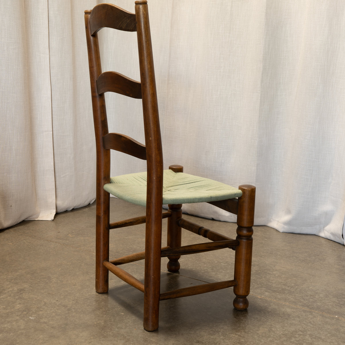 A wooden ladder-back dining chair with a light green woven seat, positioned against a draped white fabric backdrop.