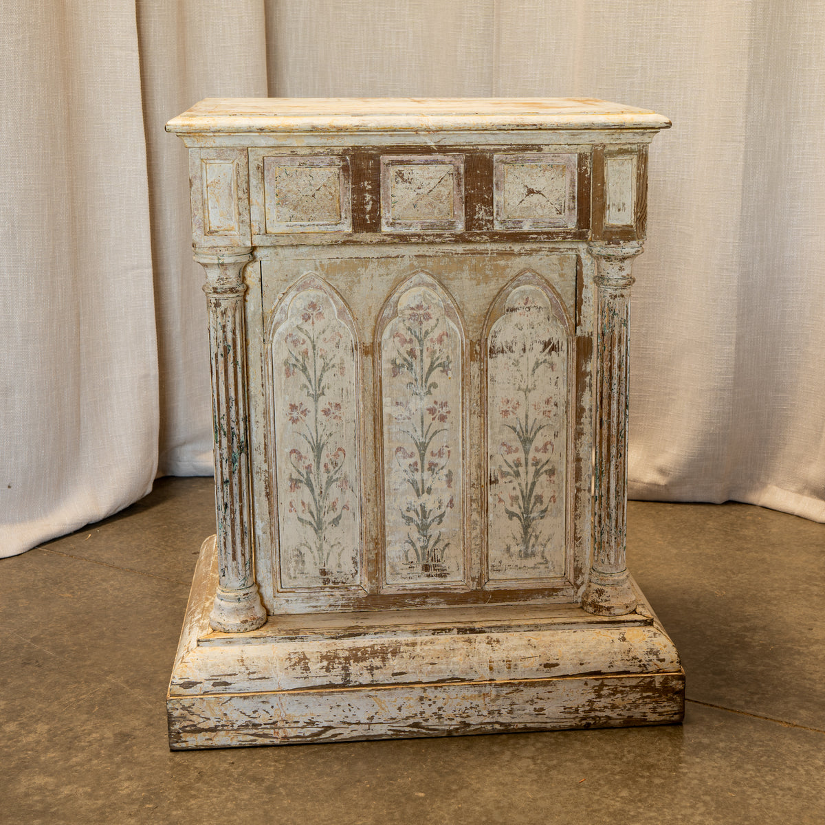 A distressed, vintage-style wooden side table or cabinet with a weathered off-white and brown paint finish, featuring three arched floral panels on the front and decorative columns on the sides, set against a neutral draped fabric backdrop.