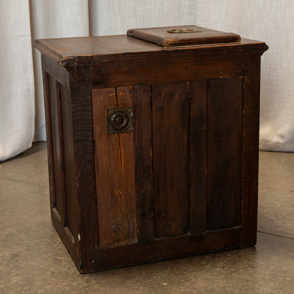 A rustic dark brown wooden chest or box with vertical plank panels, a hinged lid, and an ornate round metal knob, photographed indoors against a light fabric backdrop and concrete floor.