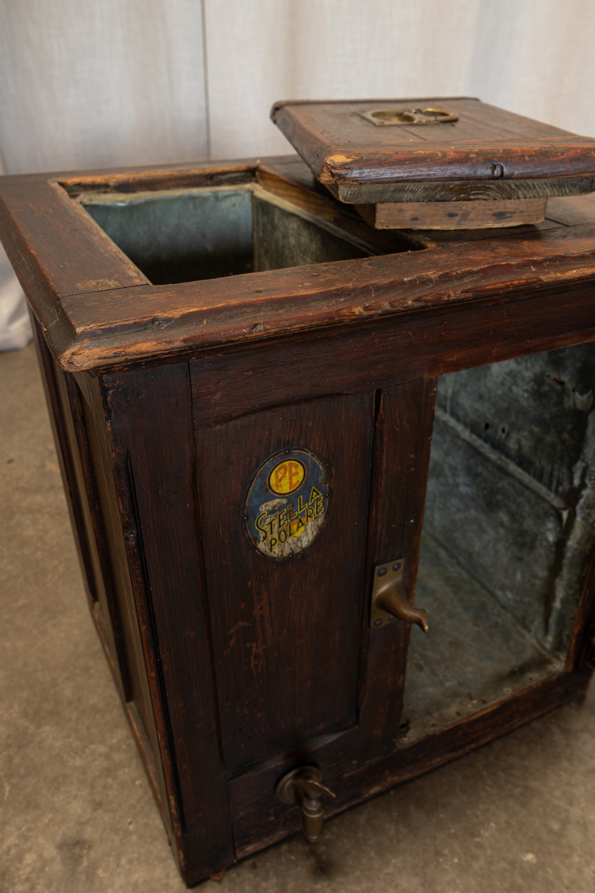 A vintage dark brown wooden liquor cabinet with a glass door, brass tap handle, and a hinged wooden lid resting on top, set against a neutral indoor backdrop.