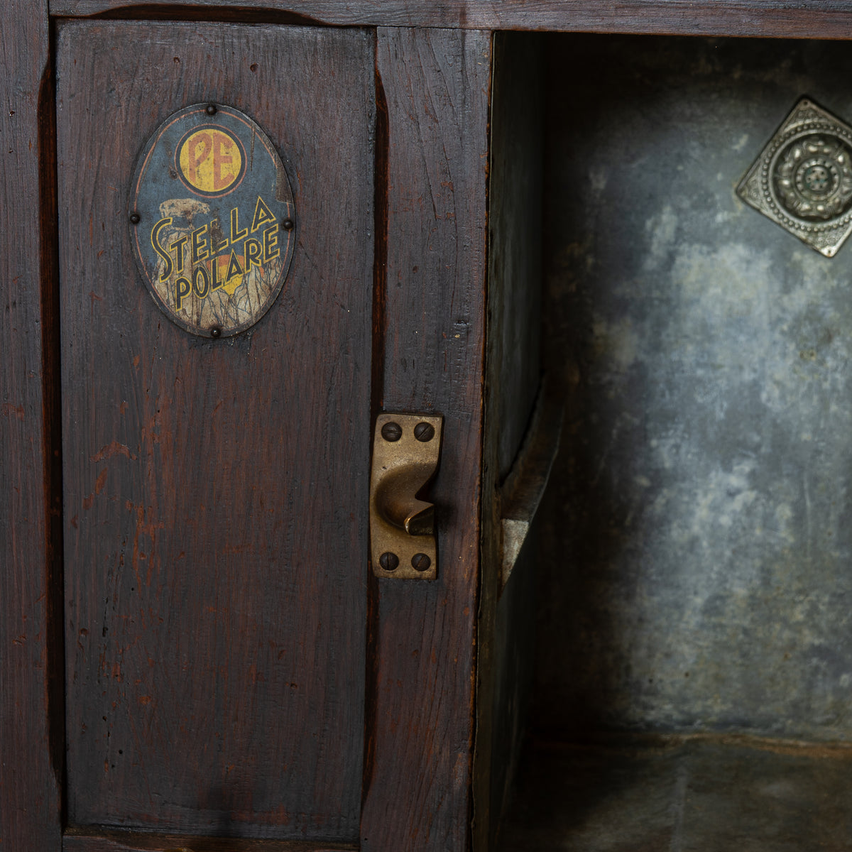 Close-up of a dark wooden cabinet door with a brass handle and a round brass plaque reading 'STELLA POLINE', next to an open compartment with a textured gray interior.
