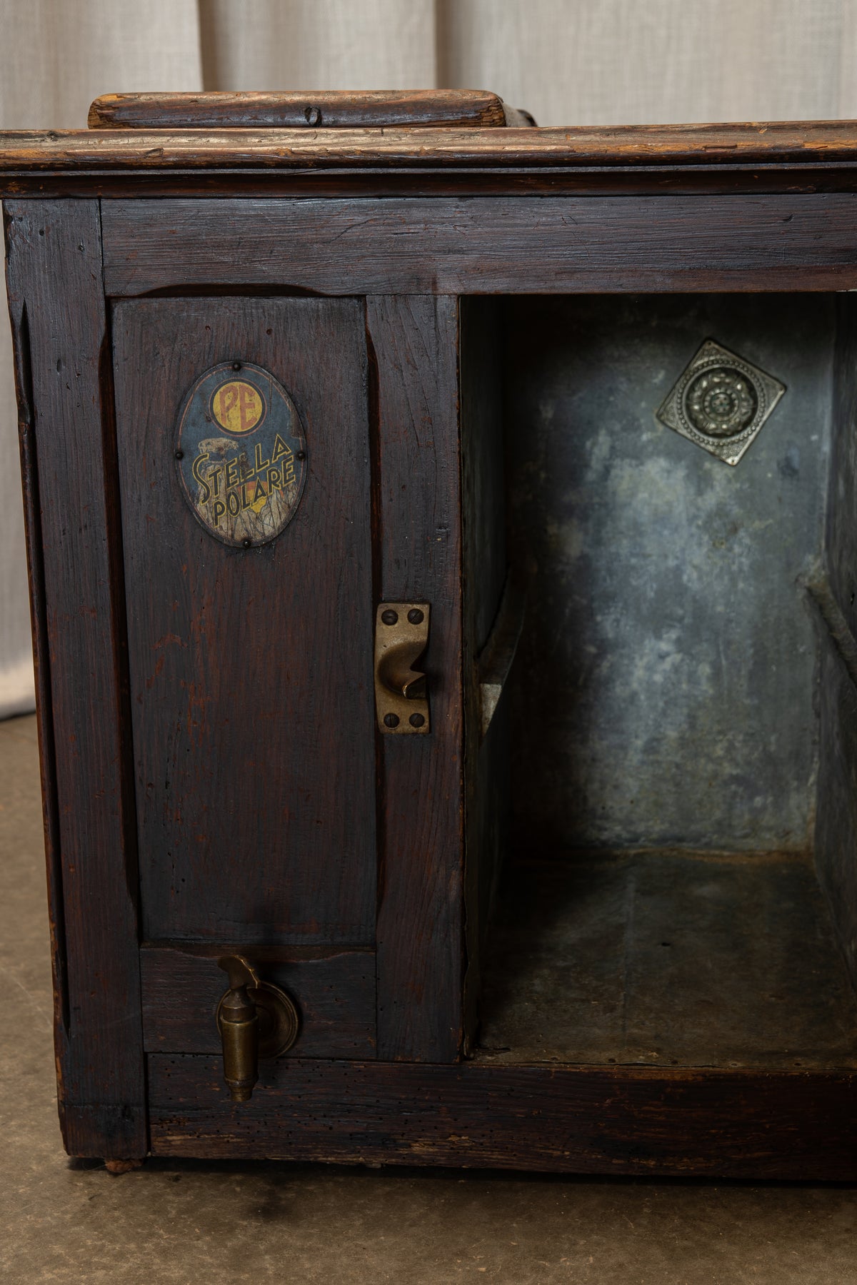 An antique dark wood chest or cabinet with a weathered finish, featuring brass hardware, a small oval decorative plaque on the door, and an open side compartment revealing a galvanized metal interior.