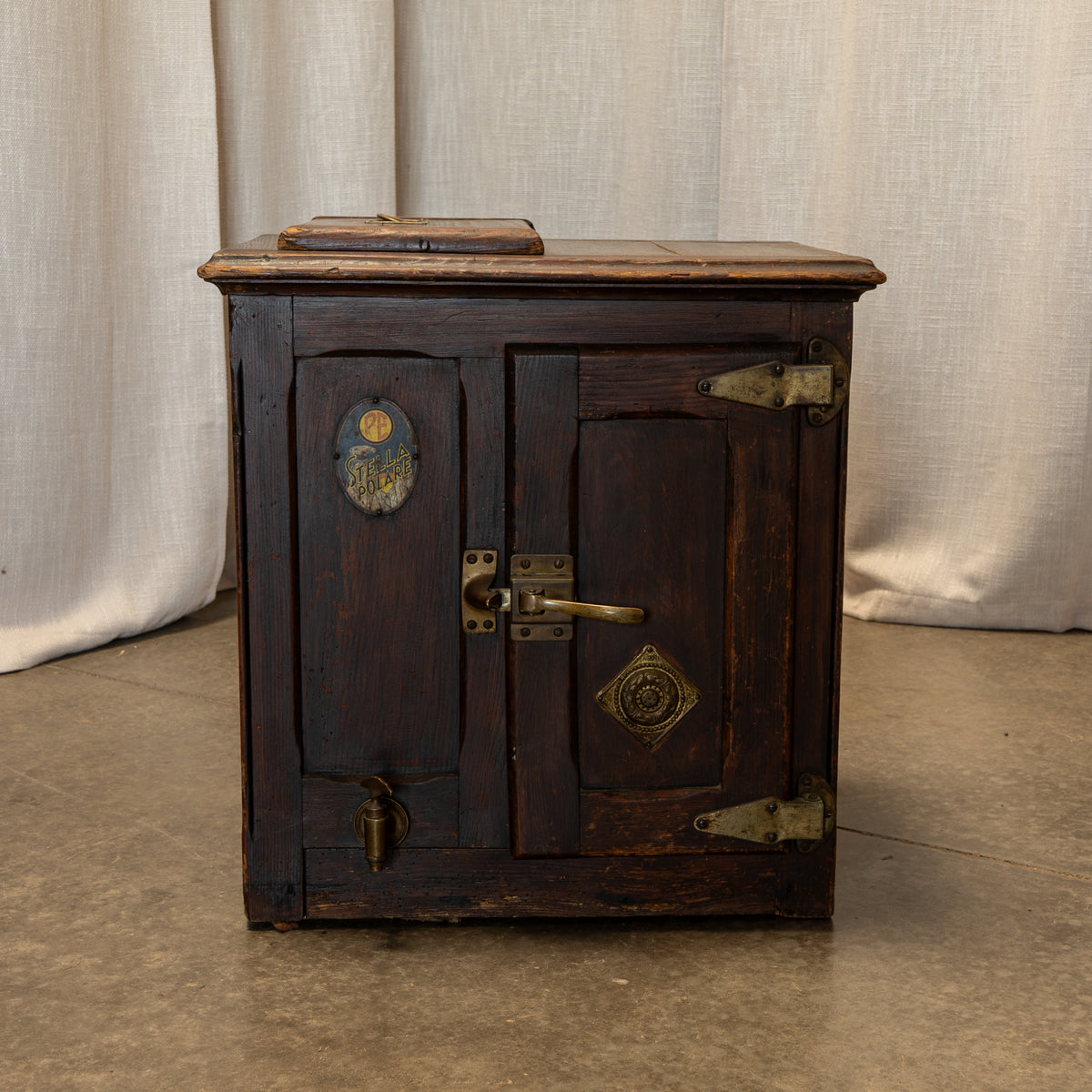 A rustic dark brown wooden chest or cabinet with distressed finish, antique brass hardware, and decorative medallions, set against a neutral beige curtain backdrop.
