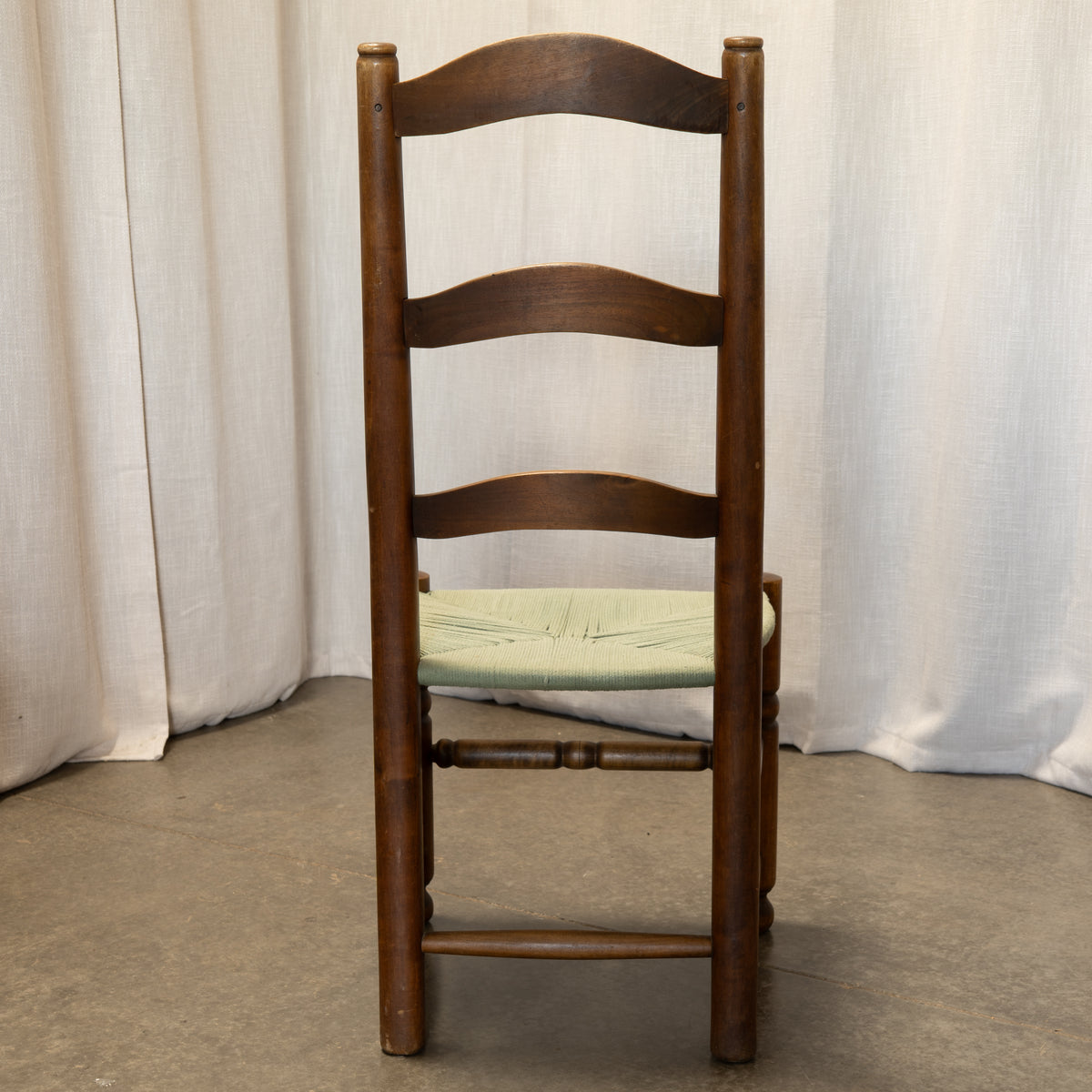 A wooden ladder-back dining chair with a light green cushioned seat, photographed against a neutral white curtain backdrop.
